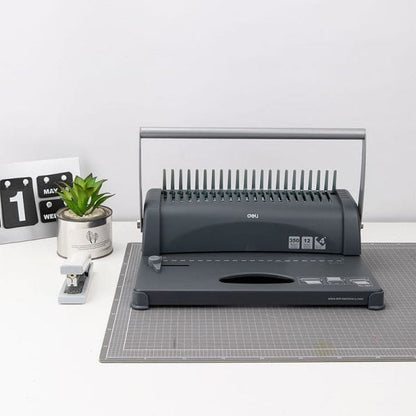 Black office binding machine on a gray cutting mat with a small plant and calendar in the background.
